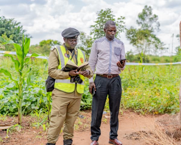 African environmental officer in safety vest and man in business attire inspecting agricultural farmland with growing crops and crime scene tape.