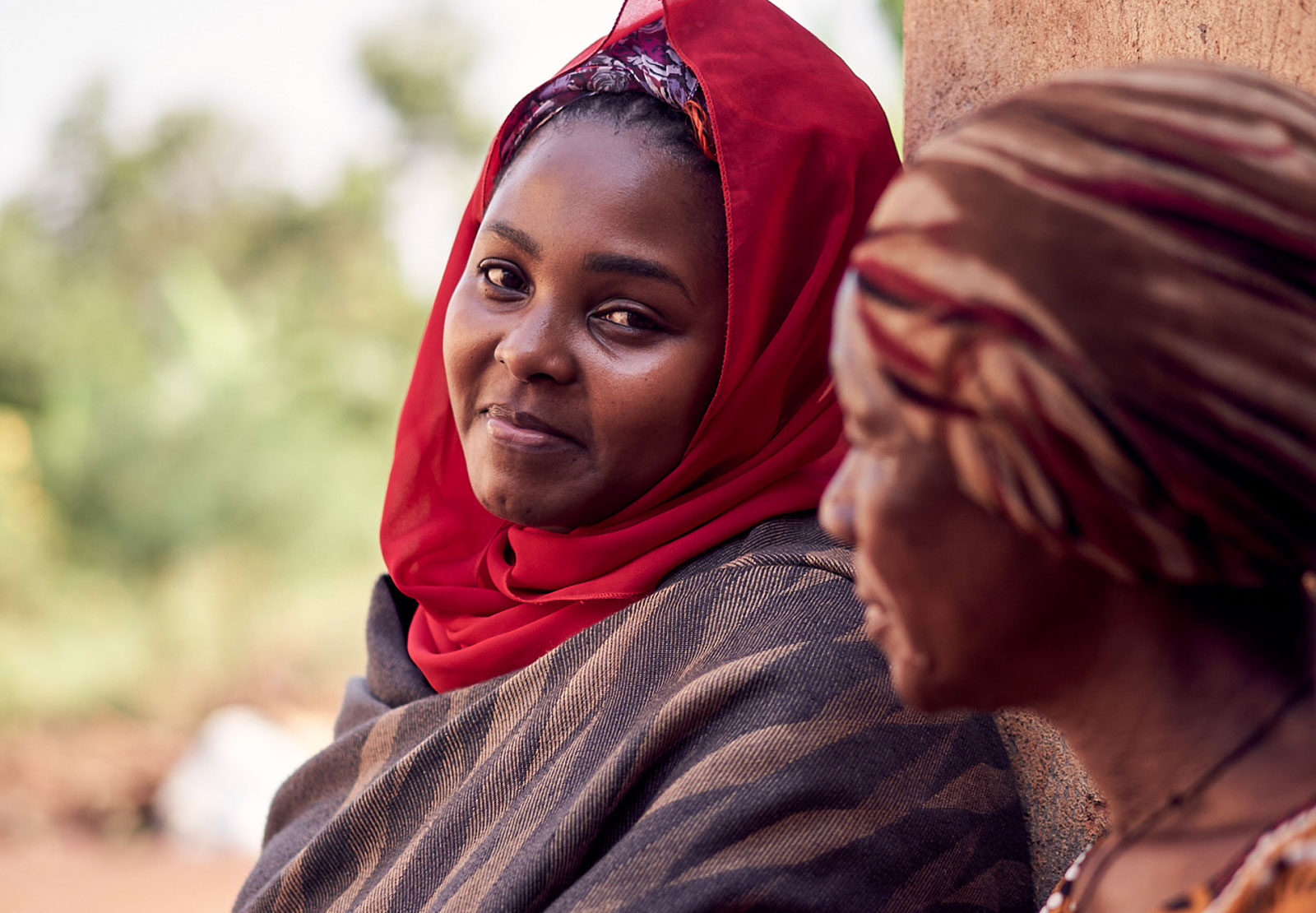 Two women reclining against a wall outdoors, one is wearing a red headscarf